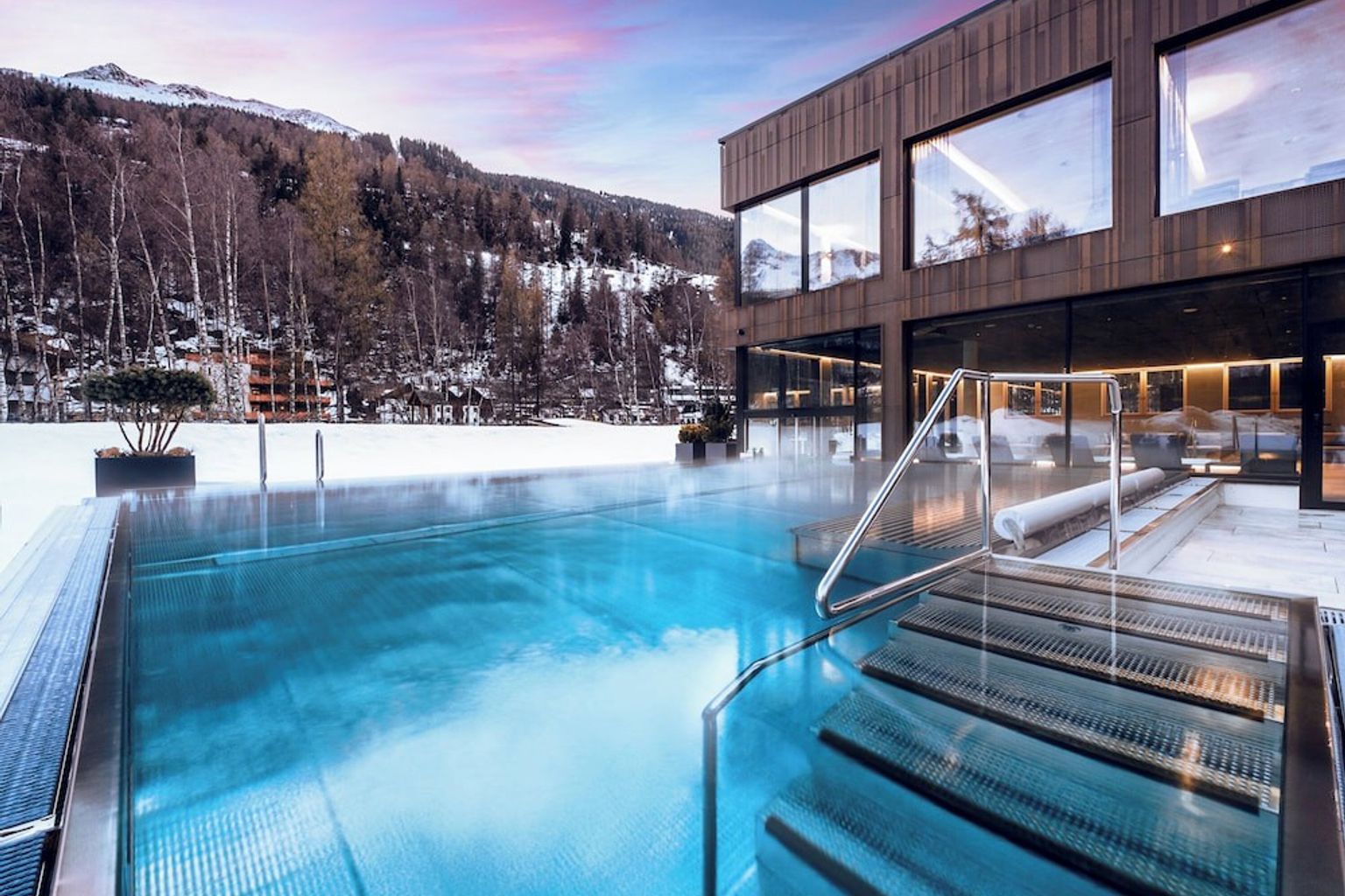 Et dampende utendørsbasseng ved en moderne bygning, omgitt av snødekte fjell.
