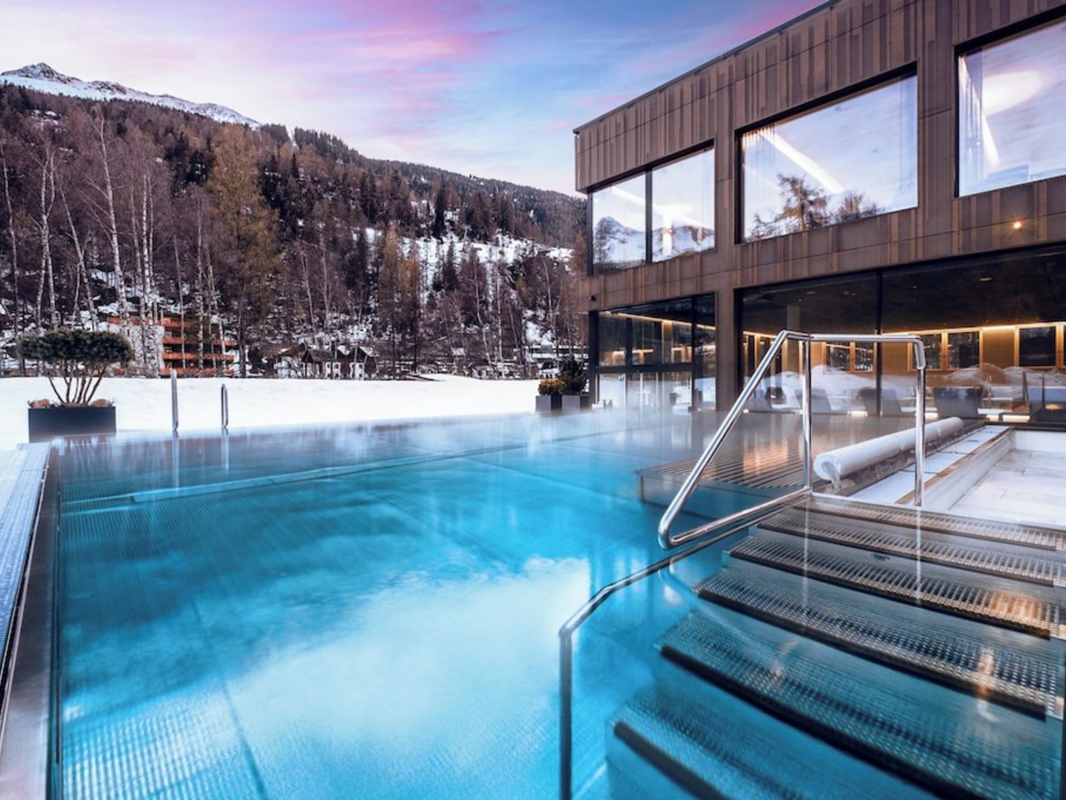 Et dampende utendørsbasseng ved en moderne bygning, omgitt av snødekte fjell.
