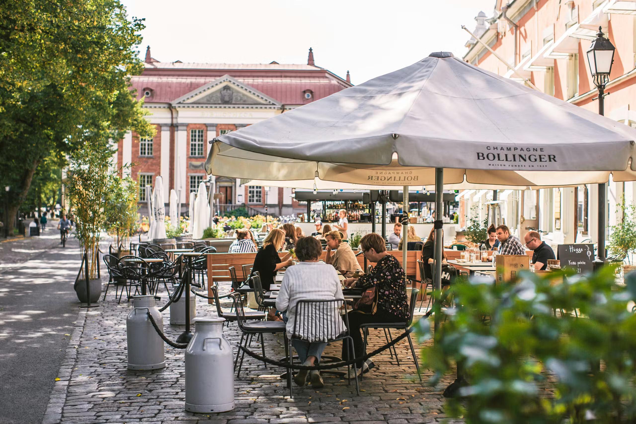 Et udendørs café med mennesker, der spiser under en Bollinger-paraply på en brostensbelagt gade, flankeret af træer og historiske bygninger.