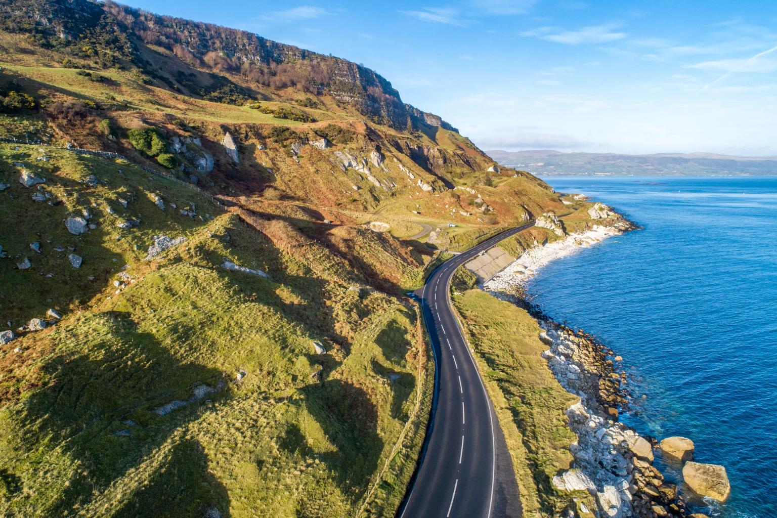 Aerial view of a winding coastal road along rocky green hills and the blue ocean.