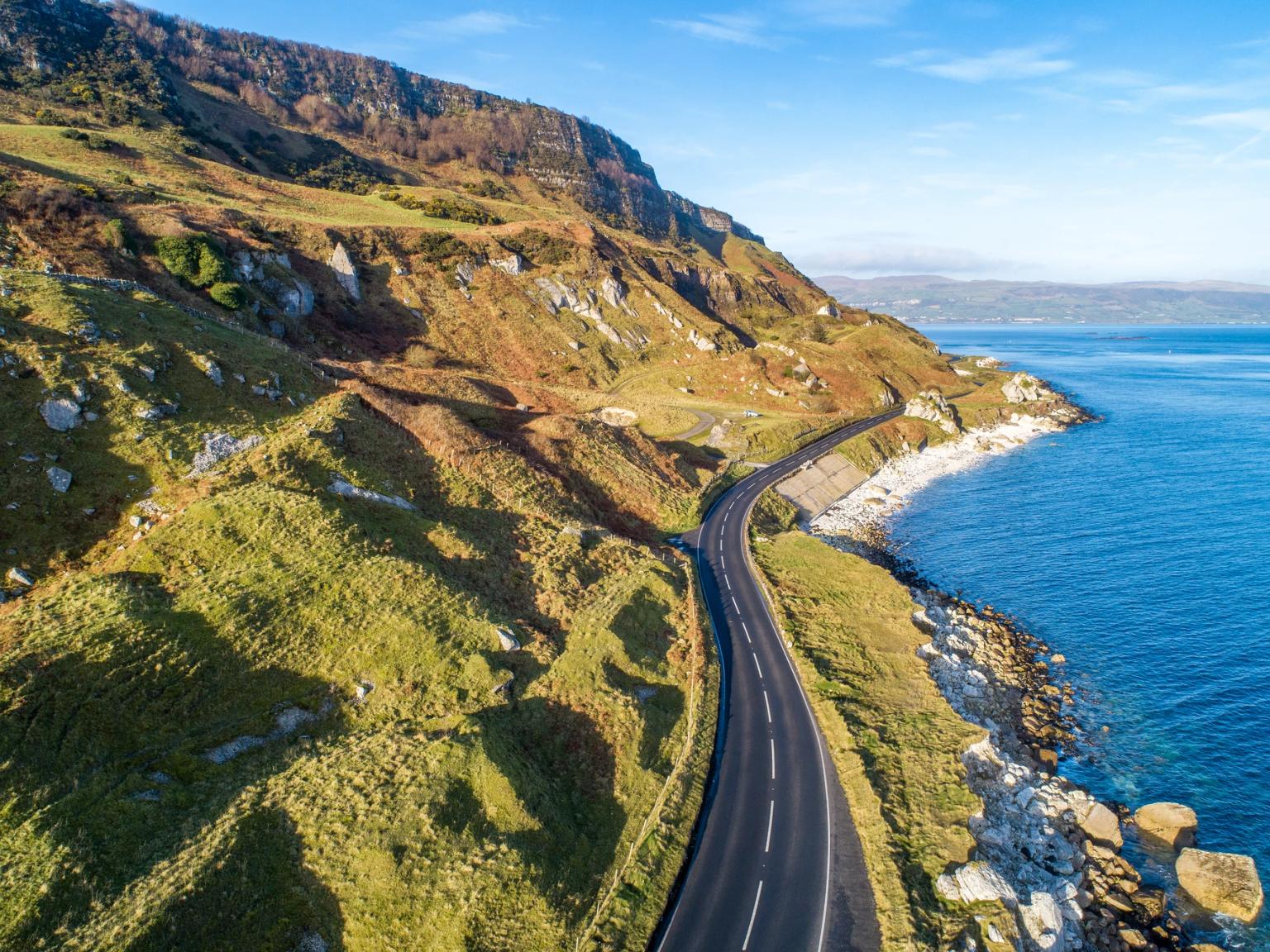 Aerial view of a winding coastal road along rocky green hills and the blue ocean.
