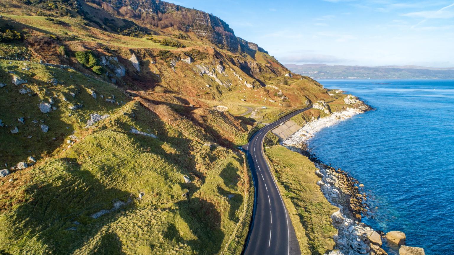 Aerial view of a winding coastal road along rocky green hills and the blue ocean.