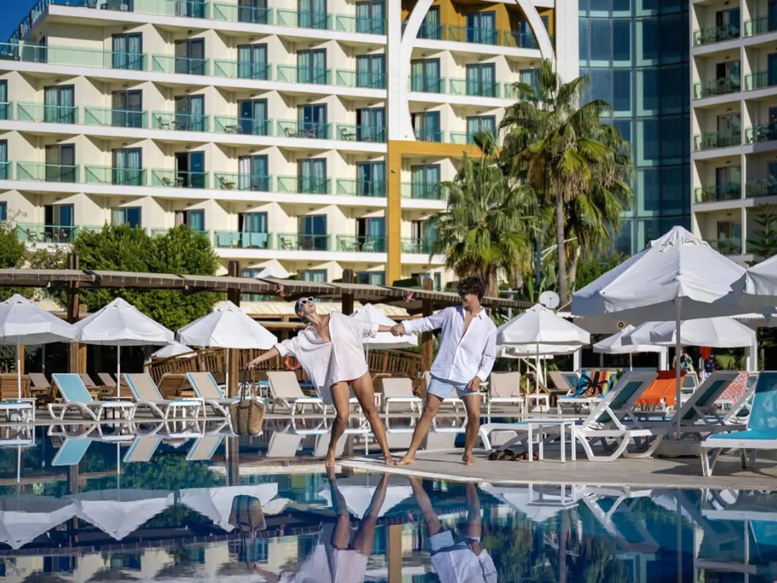 En man och kvinna i vita skjortor poserar lekfullt vid en pool med ett resorthotell och palmer i bakgrunden. Alan Xafira Deluxe Resort & Spa, Alanya