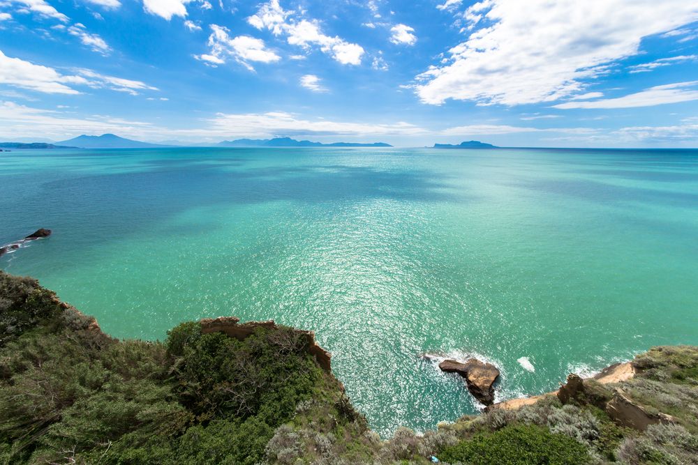 Vue en plongée sur un océan turquoise depuis des falaises rocheuses sous un ciel bleu avec des nuages, avec une terre lointaine à l’horizon. Procida, Italie