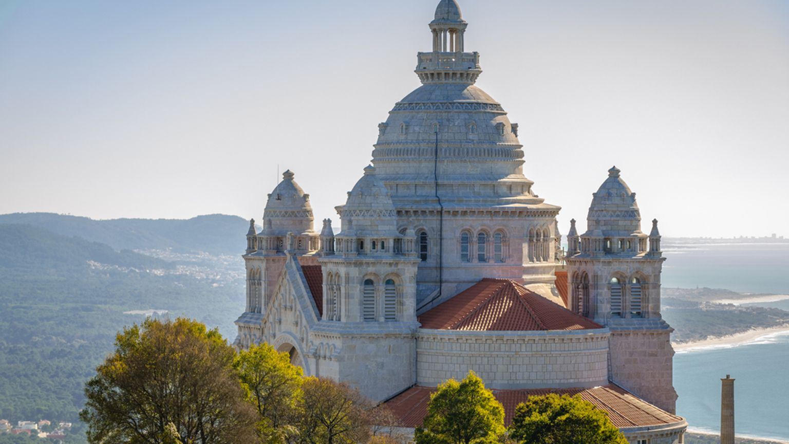 En majestätisk basilika i vit sten med en kupol och torn, belägen på en kulle ovanför bergen och havet.