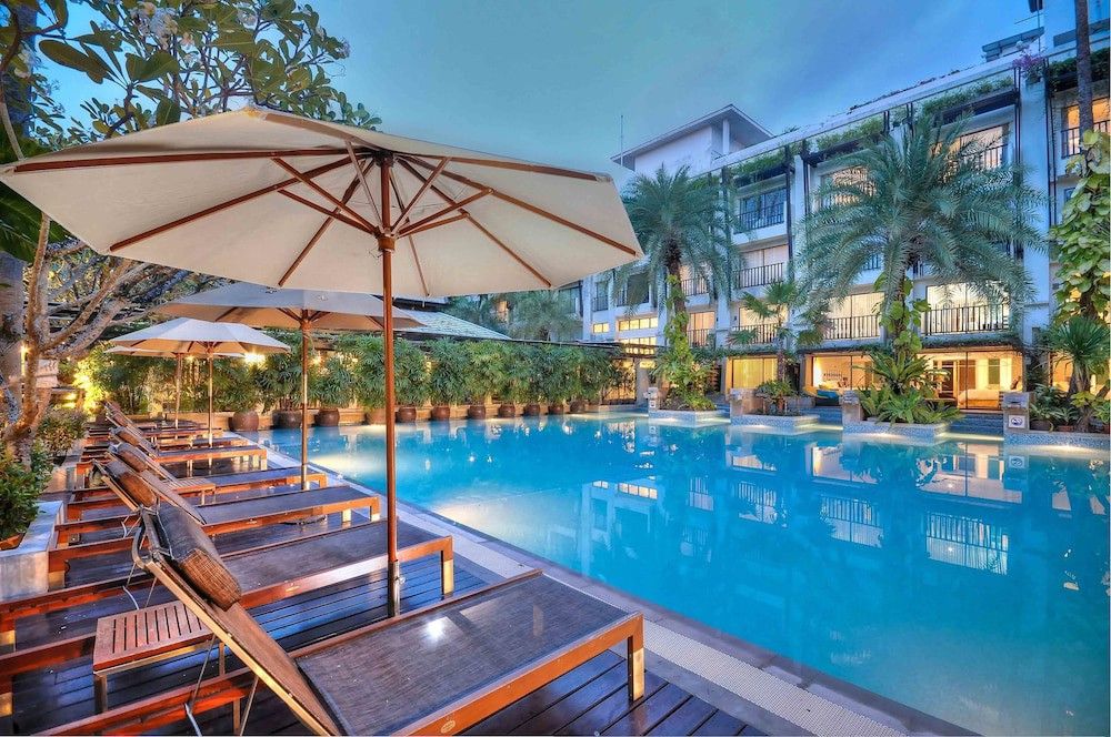 En upplyst hotellpool i skymningen, med solstolar, parasoller och en tropisk byggnad.
