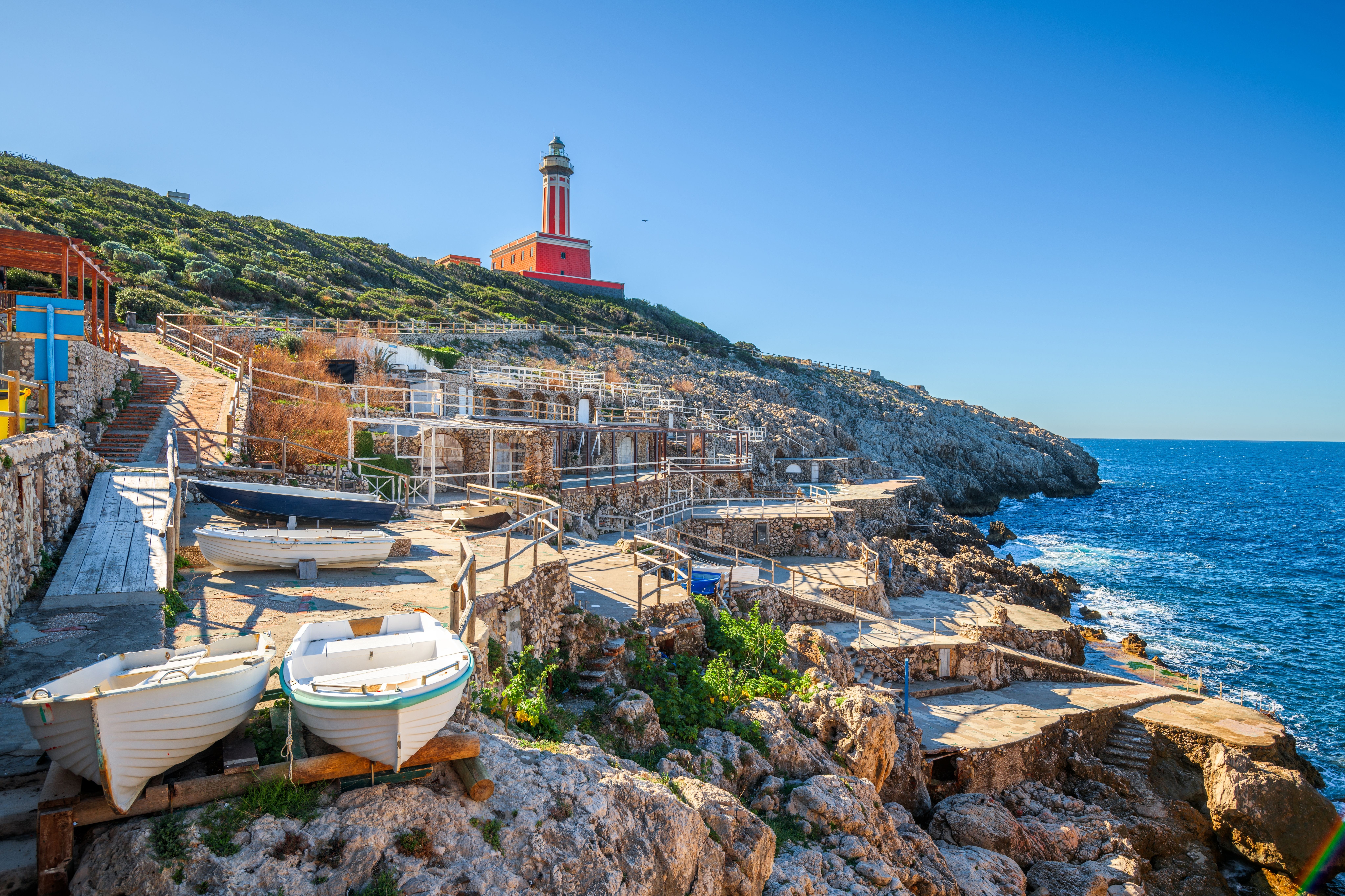 Capri. Un phare rouge sur une colline verte surplombe un littoral rocheux avec des constructions en terrasses, des bateaux et la mer bleue.