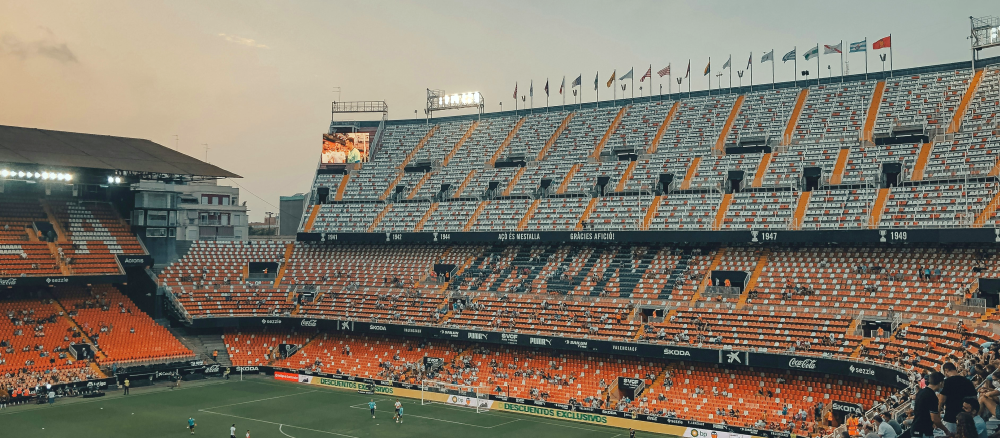 Soccer stadium with orange and white tiered seats spelling 'VALENCIA', some spectators, players on the field, and flags under an evening sky.