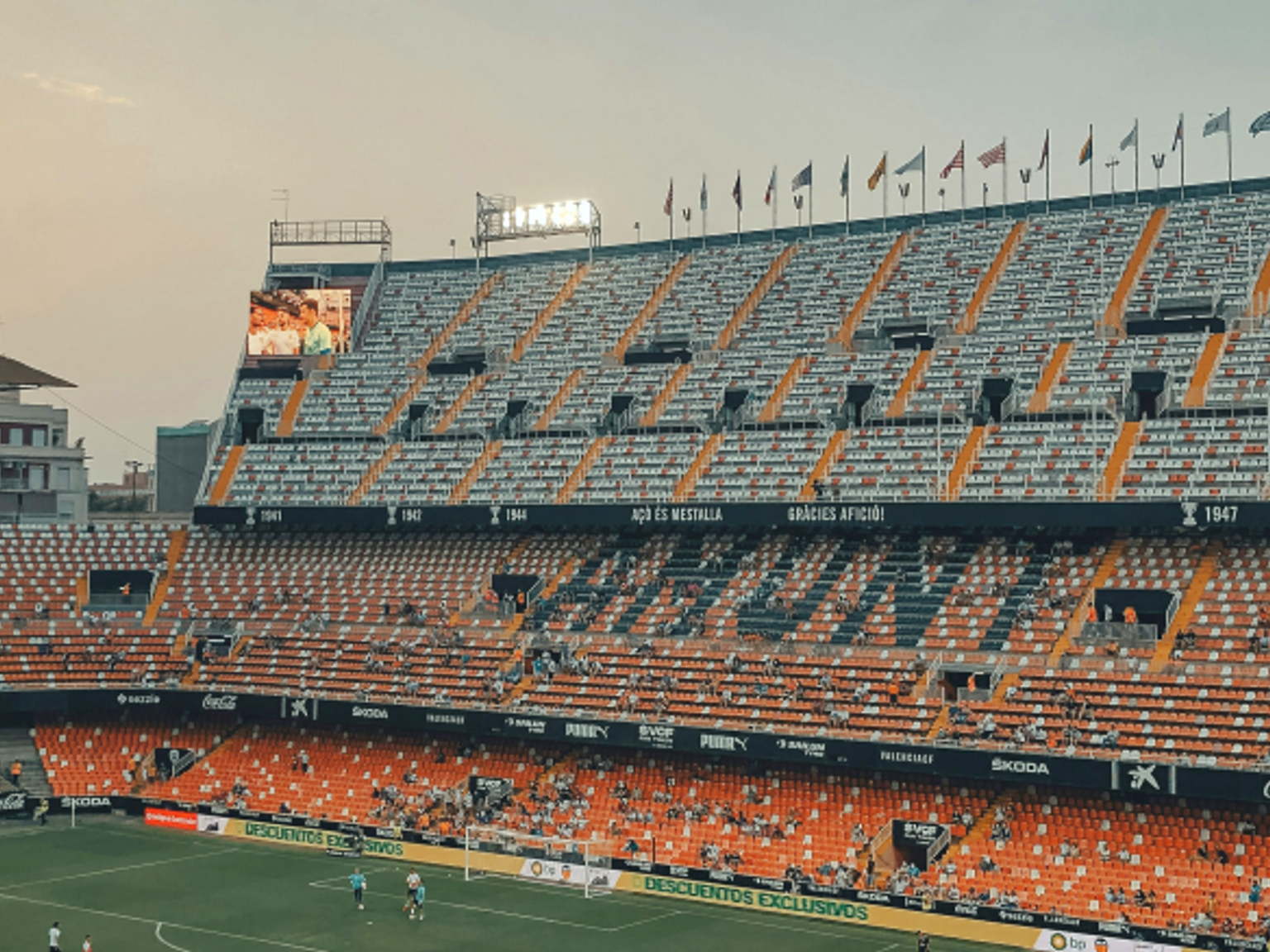 Soccer stadium with orange and white tiered seats spelling 'VALENCIA', some spectators, players on the field, and flags under an evening sky.