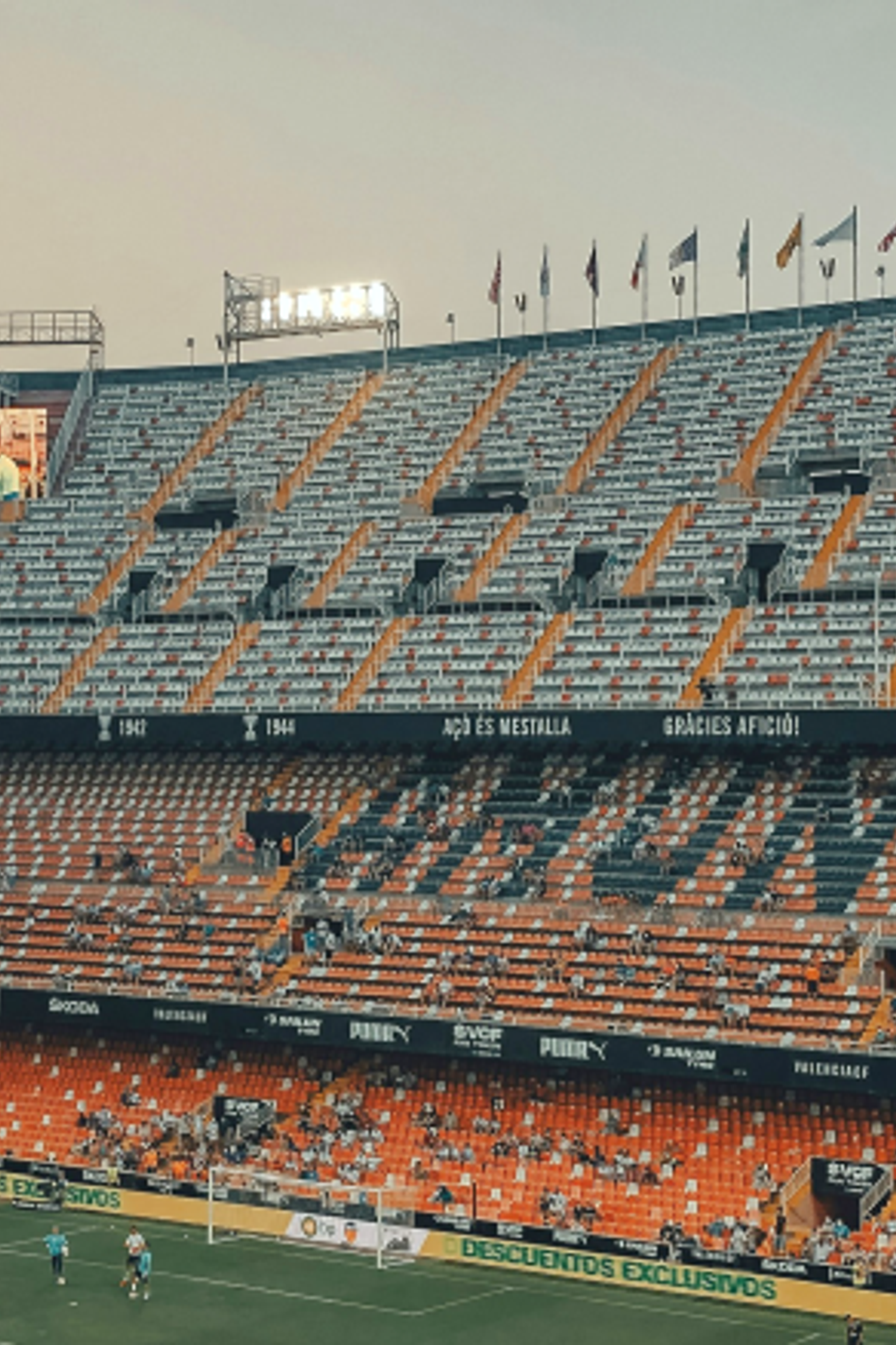 Fodboldstadion med orange og hvide trinsæder, der staver 'VALENCIA', nogle tilskuere, spillere på banen og flag under en aftenhimmel.