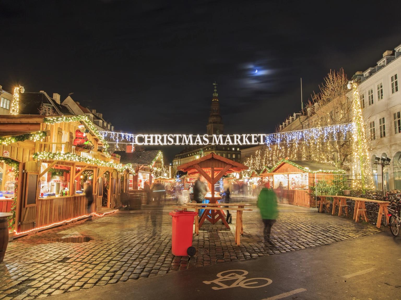 Et festligt udendørs julemarked om natten med oplyste træboder, lyskæder og et fremtrædende skilt med teksten 'CHRISTMAS MARKET'.