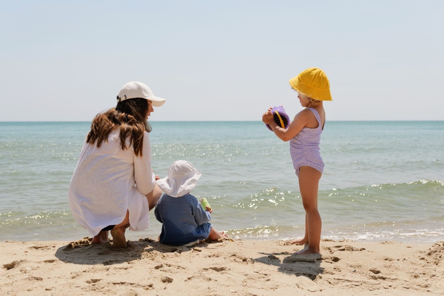 En mamma och två små barn leker på en sandstrand vid havet.