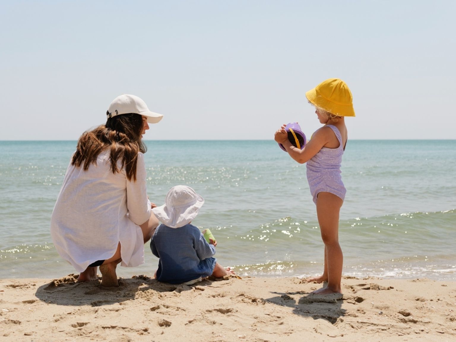 En mamma och två små barn leker på en sandstrand vid havet.