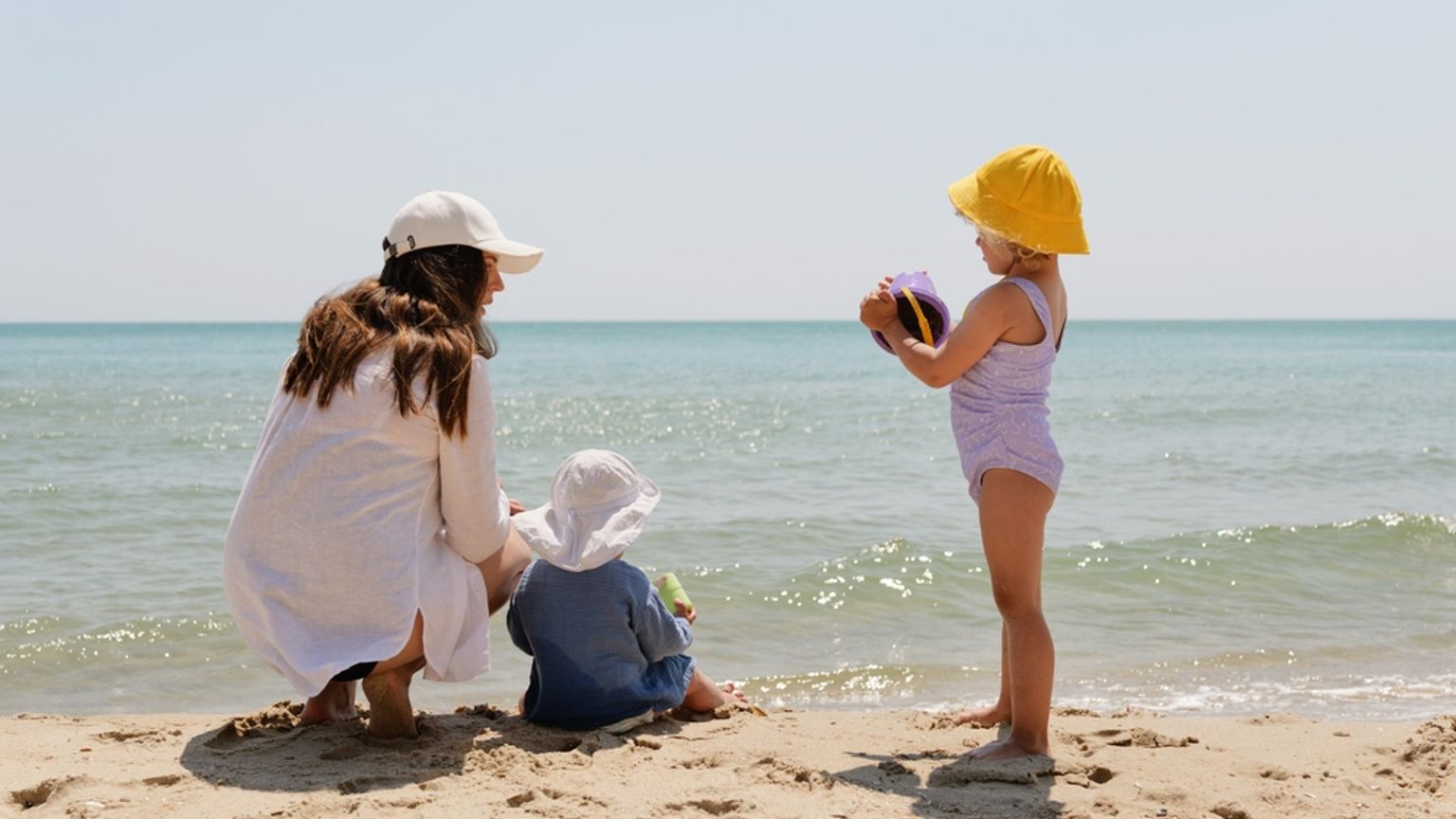 En mamma och två små barn leker på en sandstrand vid havet.