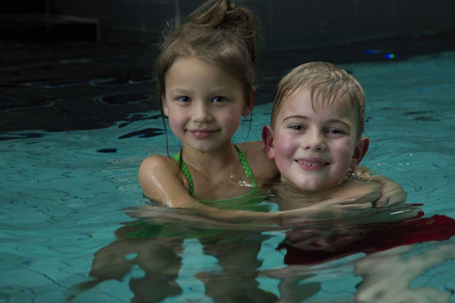 En smilende pige i en grøn badedragt og en dreng med armen omkring hende, begge i en swimmingpool.