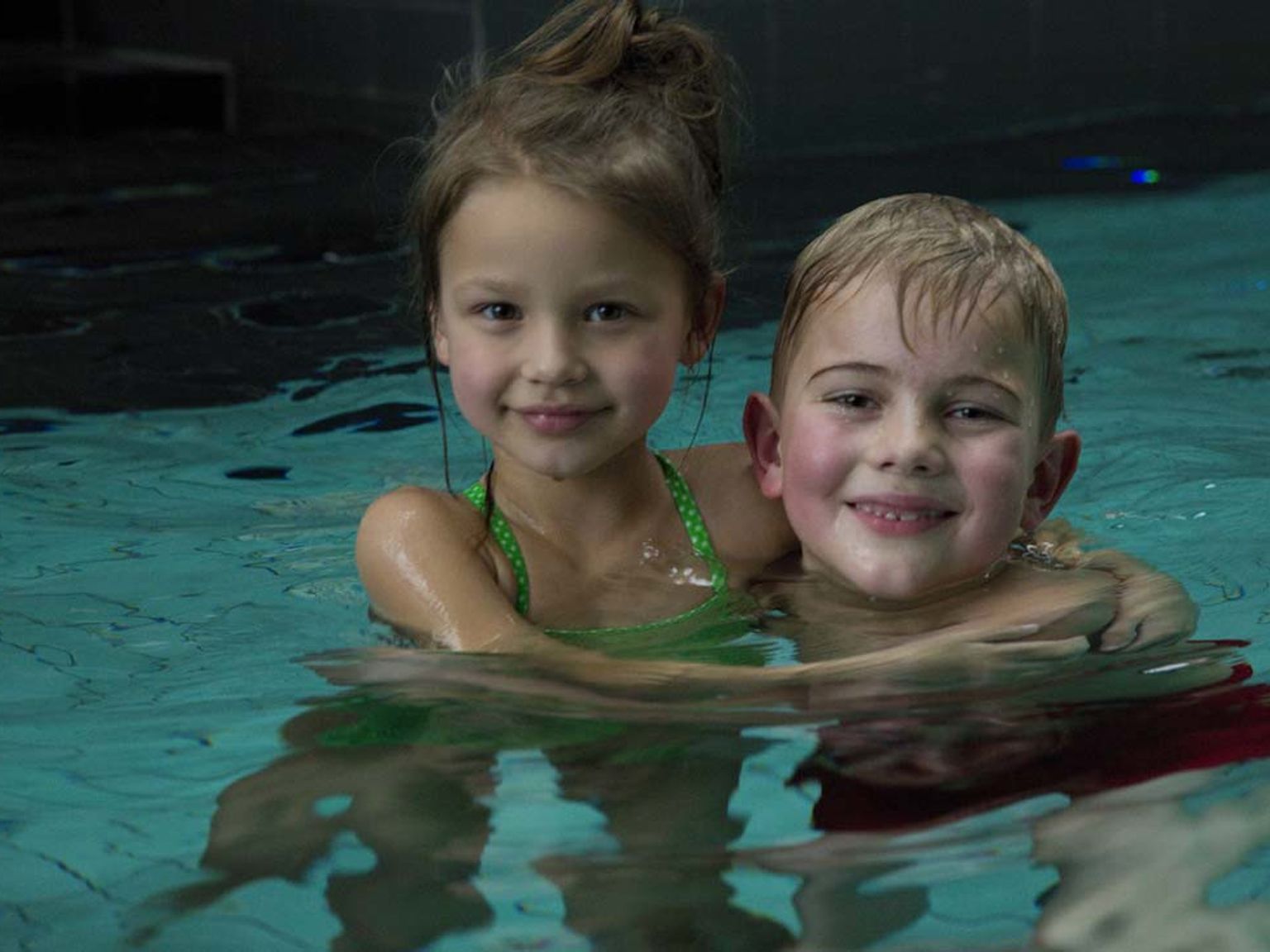 En leende flicka i en grön baddräkt och en pojke med armen runt henne, båda i en simbassäng.