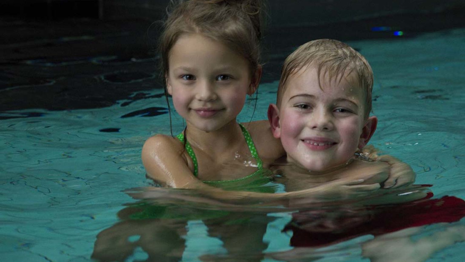 En smilende pige i en grøn badedragt og en dreng med armen omkring hende, begge i en swimmingpool.