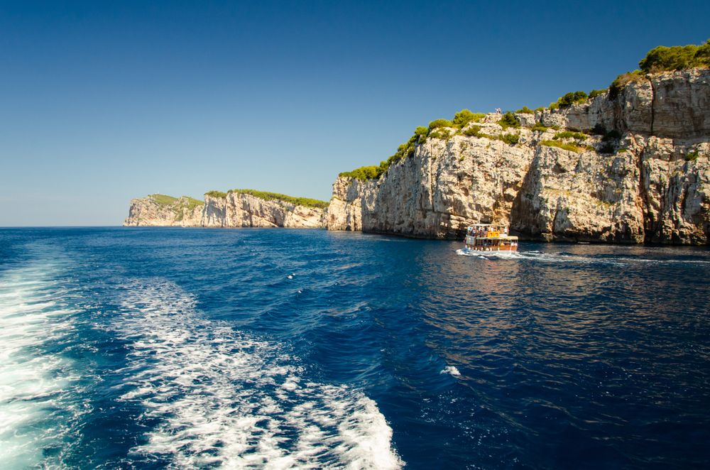 En båds kølvand på dybt blåt vand, med en turistbåd der passerer høje klipper under en klar himmel. Kornati-øerne, Kroatien