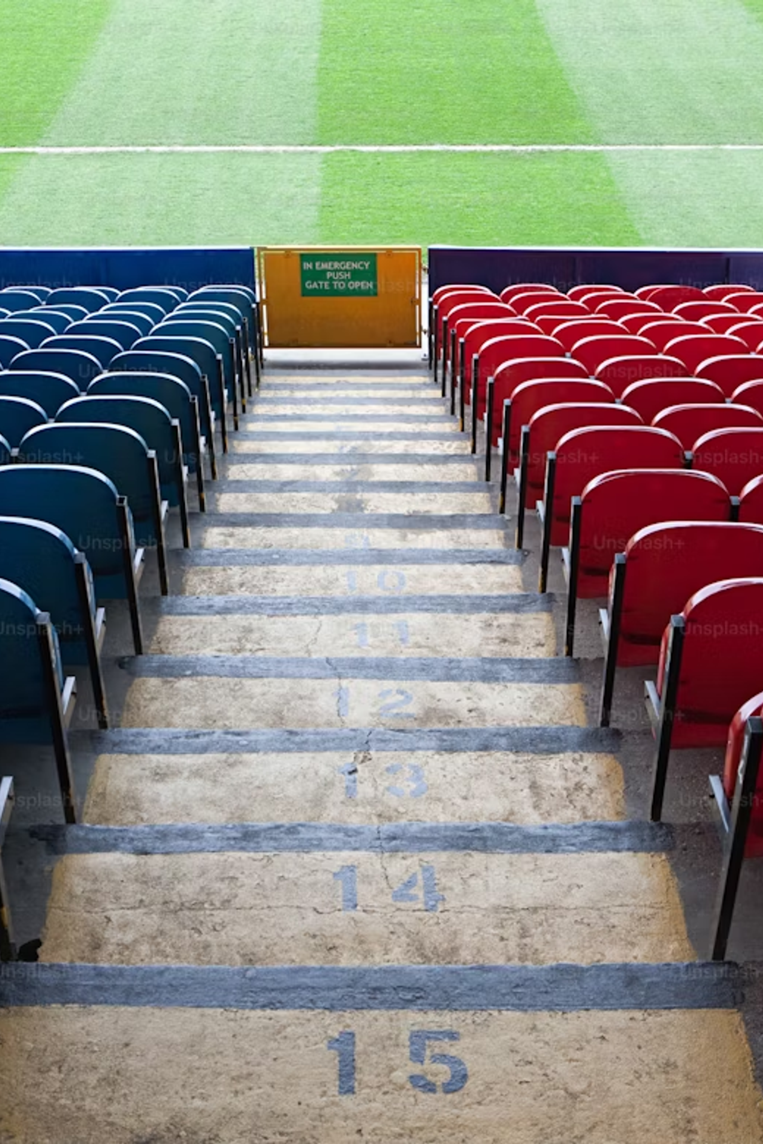 Empty blue and red stadium seats with a central aisle of stairs leading to a green pitch.