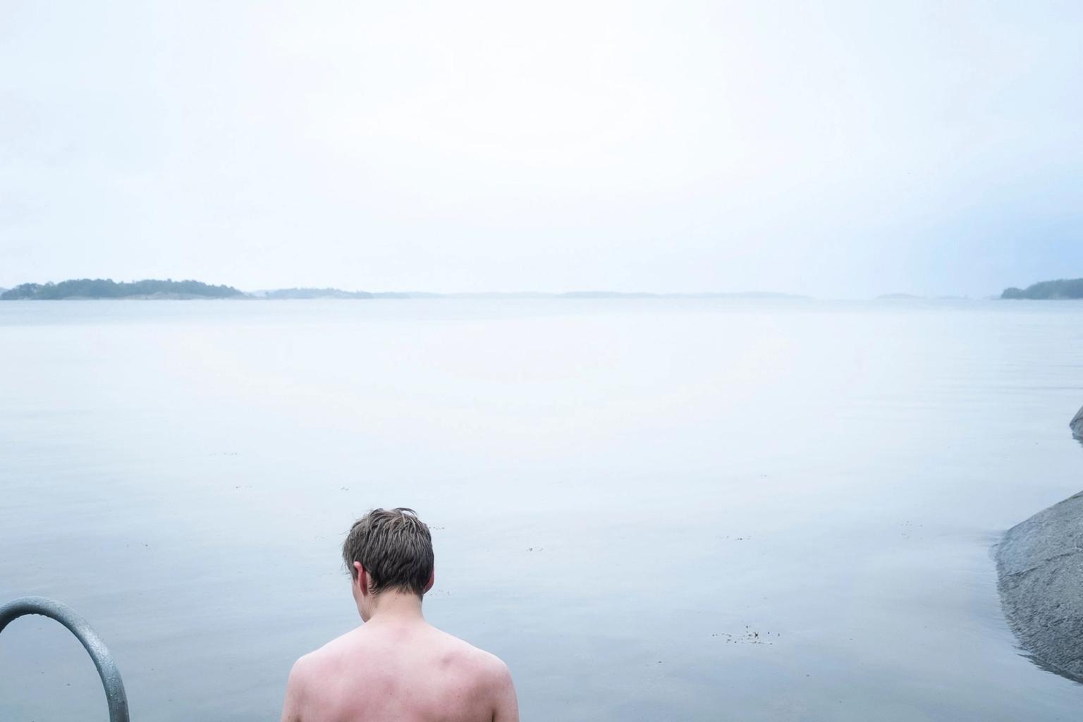 En skjorteløs mand set bagfra kigger ud over et roligt, tåget vandområde med en metalstige og fjerne øer.
