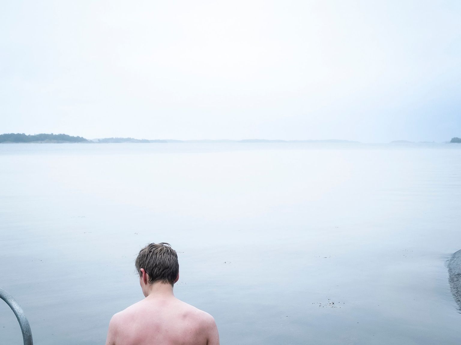 En skjorteløs mand set bagfra kigger ud over et roligt, tåget vandområde med en metalstige og fjerne øer.