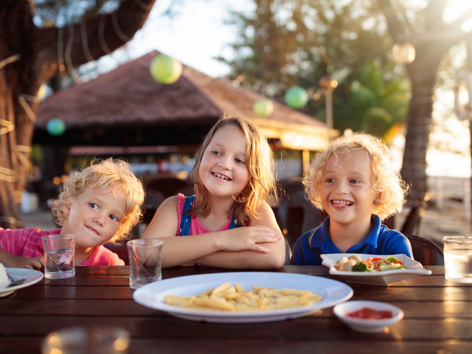 Tre glade barn sitter ved et utendørs spisebord med mat, badet i varmt solnedgangslys.