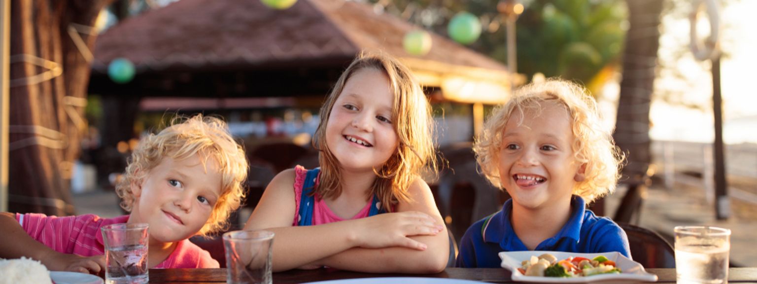 Tre glade barn sitter ved et utendørs spisebord med mat, badet i varmt solnedgangslys.