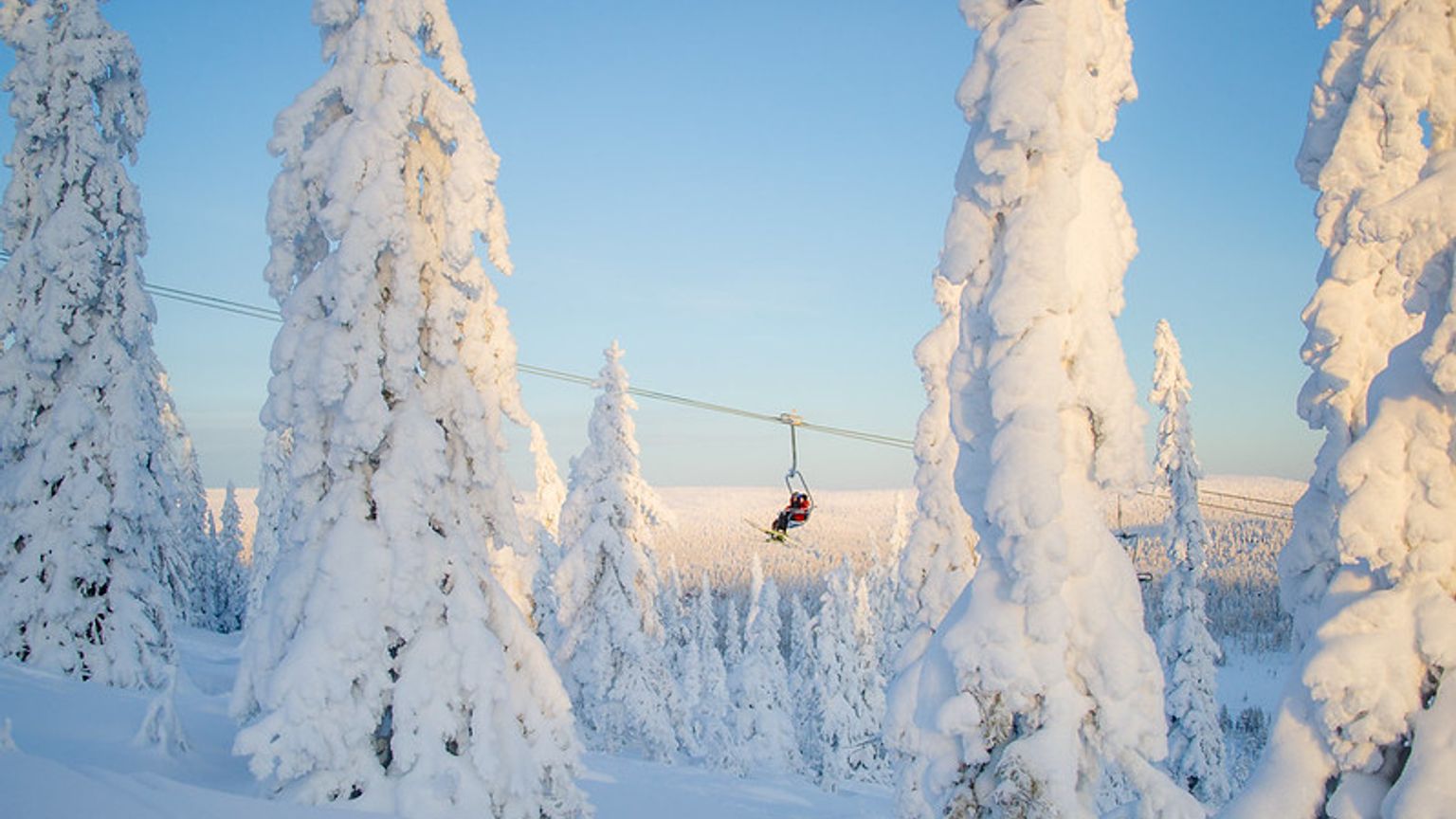 En skiløber på en stolelift stiger op over en vidstrakt, sneklædt skov af rimdækkede træer under en blå himmel.