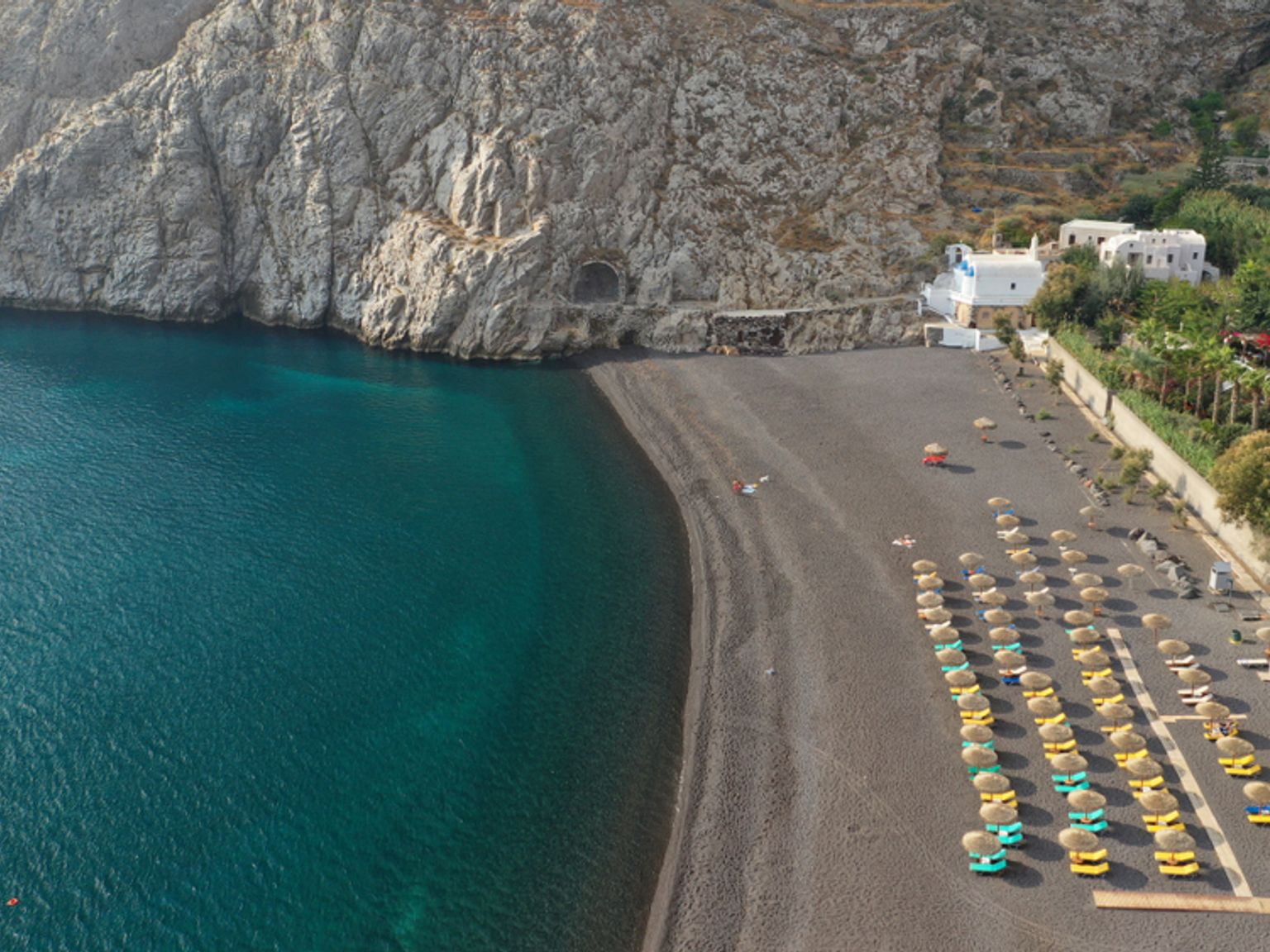 Luftfoto av en svart sandstrand med turkis hav, steinete klipper, rader med fargerike strandstoler og hvite bygninger på en åsside. Kamari, Santorini