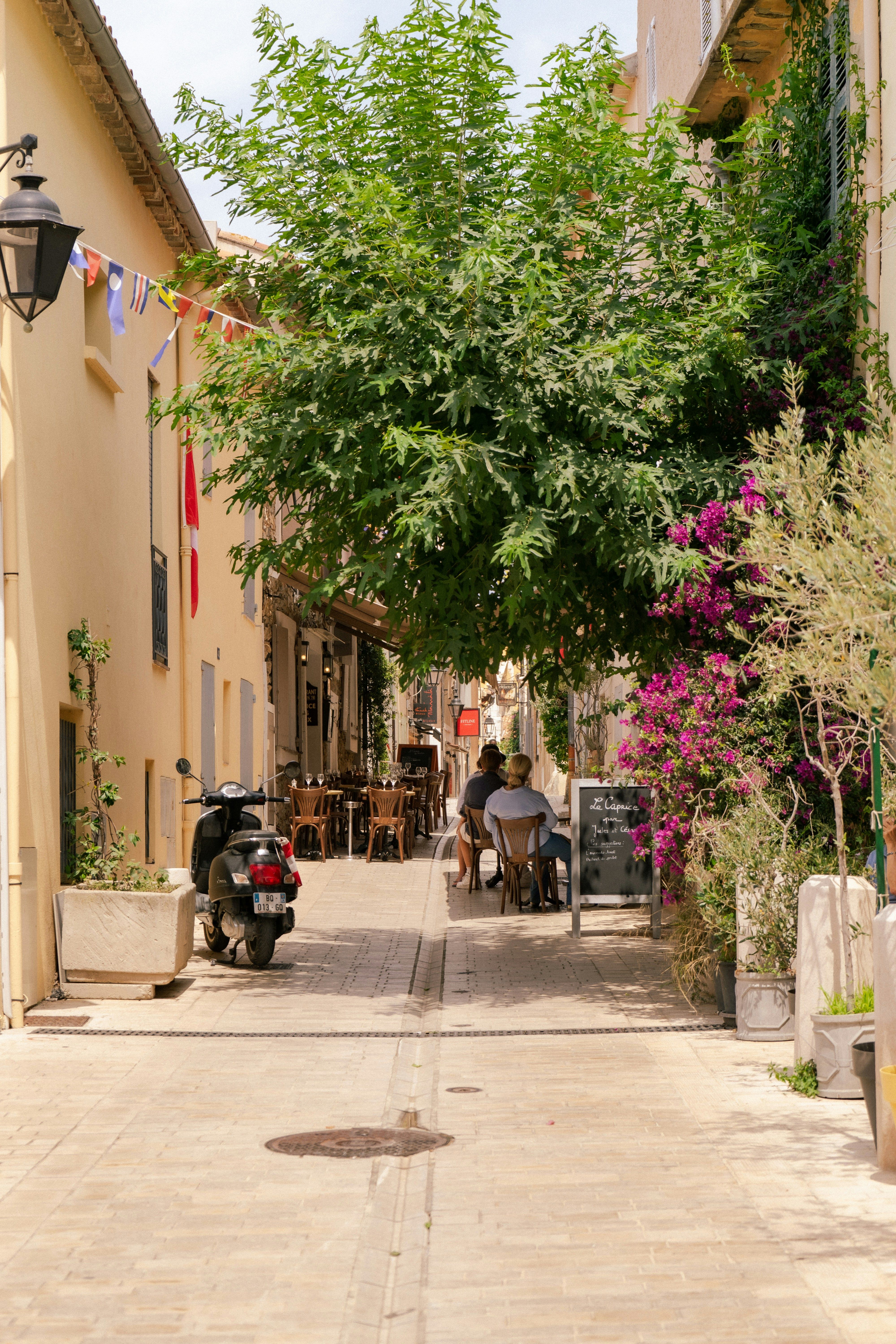 A narrow, tree-shaded street with an outdoor café, pink bougainvillea, people seated, and a scooter parked on the left. Saint-Tropez, France.