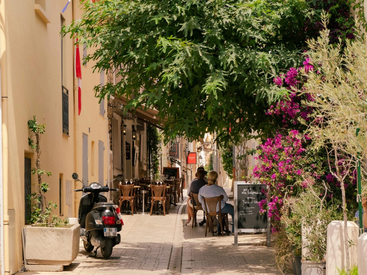 Une rue étroite ombragée par des arbres avec un café en plein air, des bougainvilliers roses, des personnes assises et un scooter garé sur la gauche. Saint-Tropez, France.