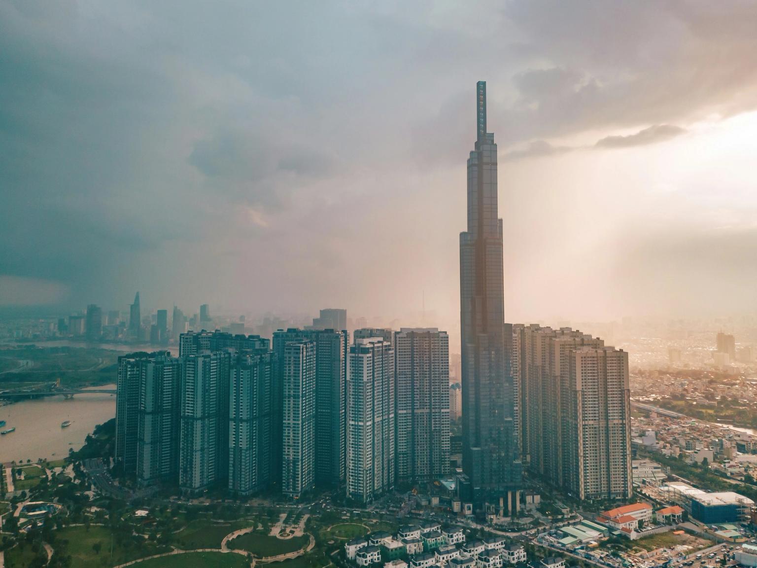 Luftfoto af en moderne by med en tårnhøj skyskraber, mange beboelsesbygninger, en flod og en diset himmel. Ho Chi Minh City