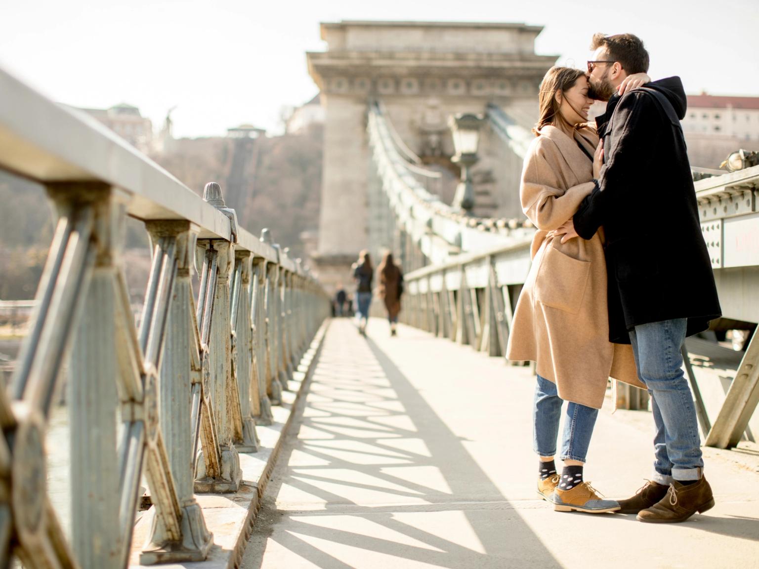 Mężczyzna całuje kobietę w czoło na słonecznym moście. Budapeszt