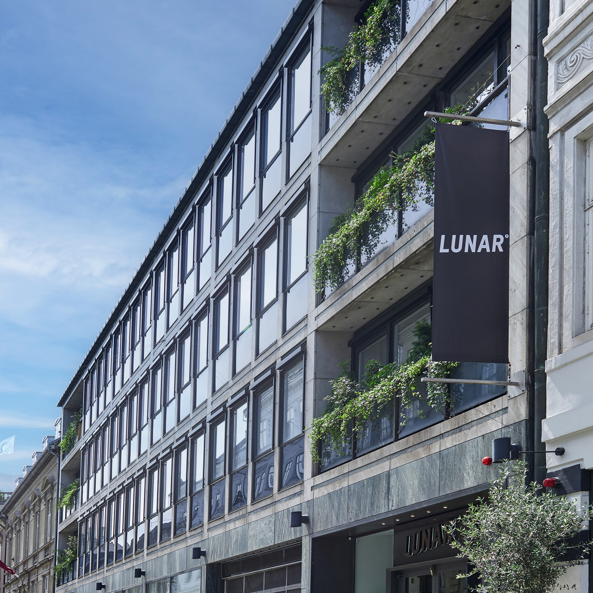 The exterior of a modern office building on a city street, with a black banner displaying the "LUNAR" logo. The facade features glass and concrete balconies with lush greenery.