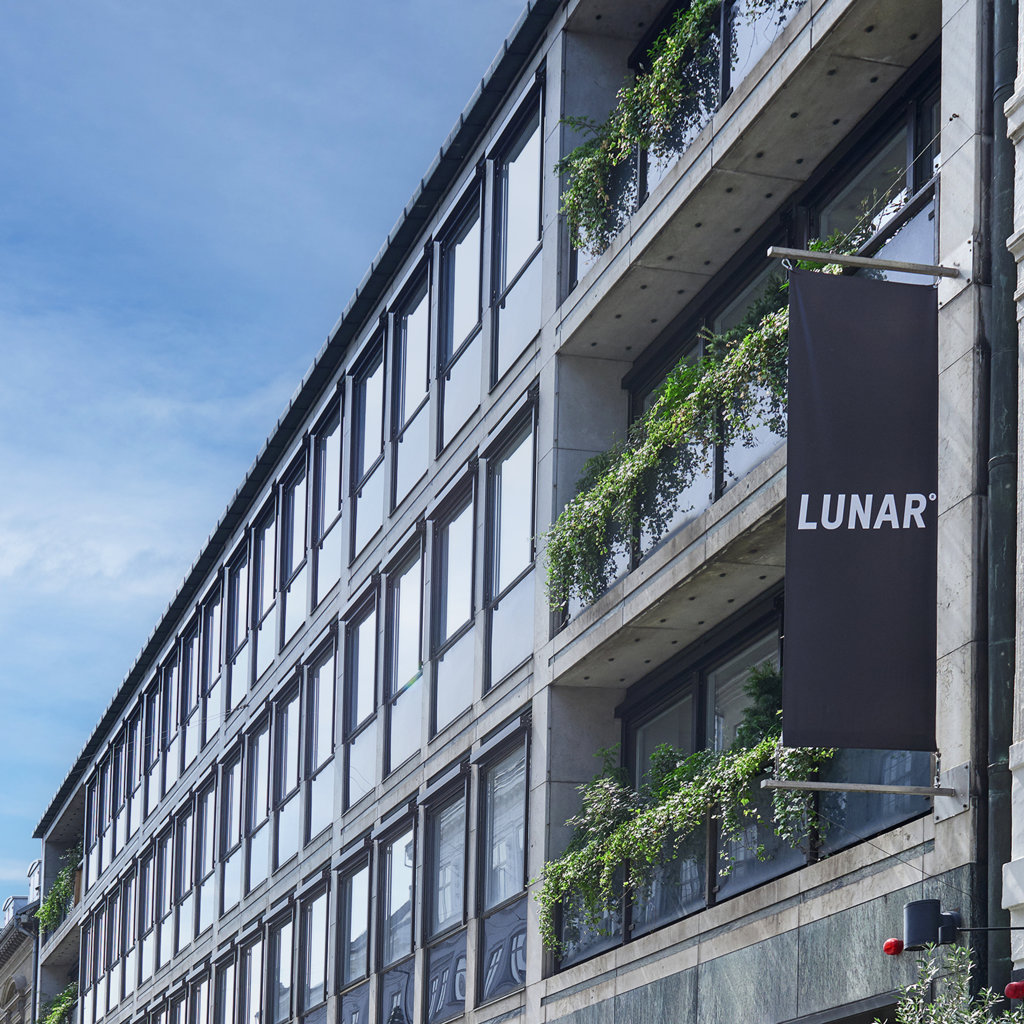 Ydersiden af en moderne kontorbygning på en bygade, med et sort banner, der viser "LUNAR"-logoet. Facaden har glas- og betonaltaner med frodige, grønne planter.