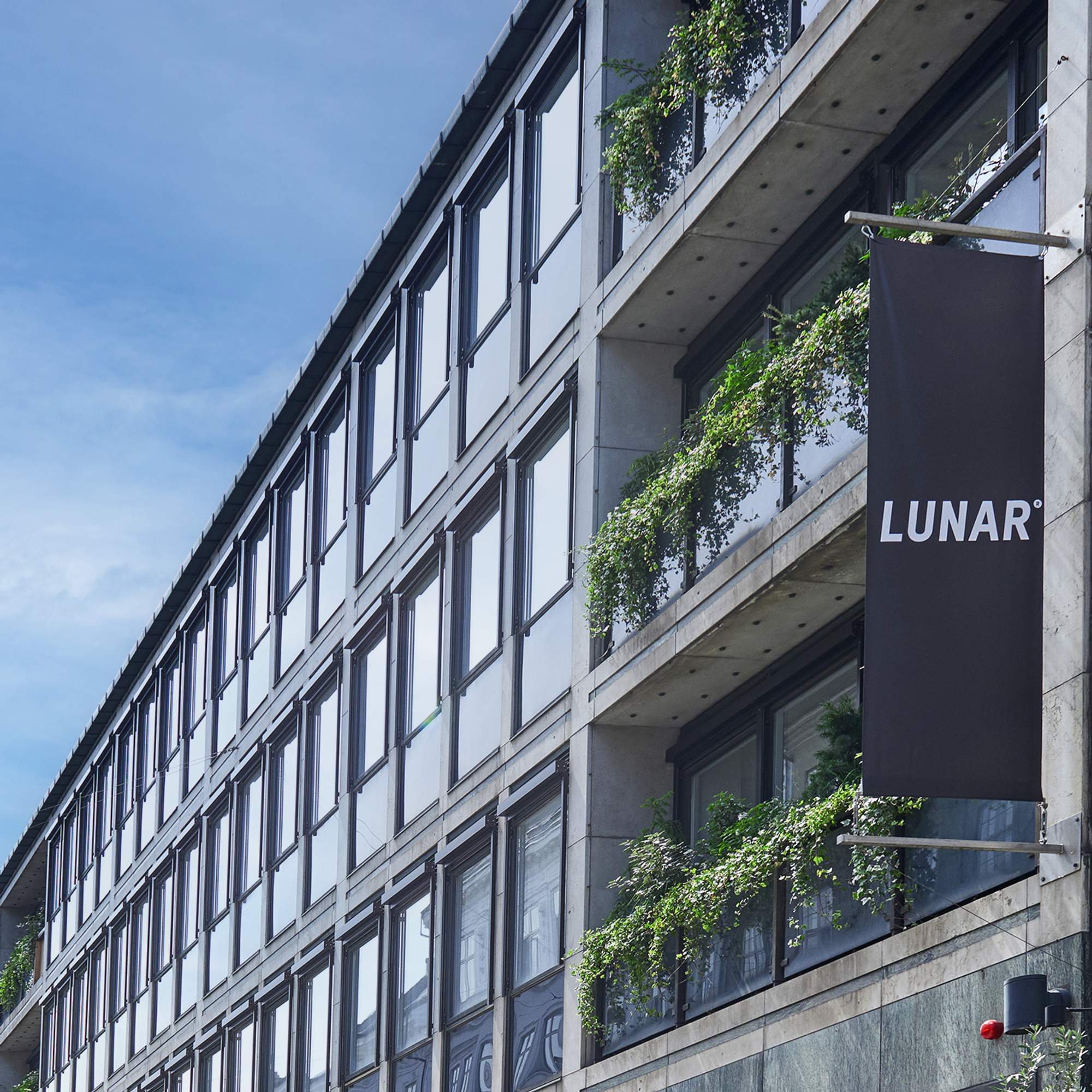 Exteriören av en modern kontorsbyggnad på en stadsgata, med en svart banderoll som visar "LUNAR"-logotypen. Fasaden har glas- och betongbalkonger med frodig grönska.