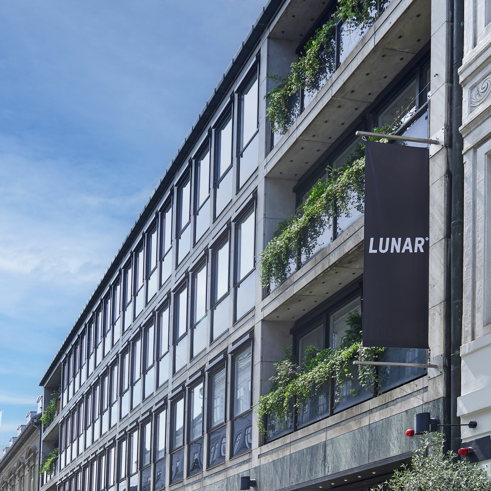 The exterior of a modern office building on a city street, with a black banner displaying the "LUNAR" logo. The facade features glass and concrete balconies with lush greenery.