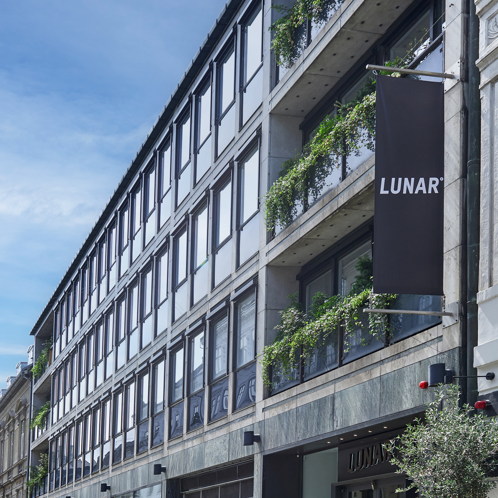 Exteriören av en modern kontorsbyggnad på en stadsgata, med en svart banderoll som visar "LUNAR"-logotypen. Fasaden har glas- och betongbalkonger med frodig grönska.