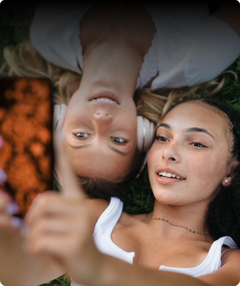 Två unga kvinnor ligger på gräset, tittar upp och ler medan de tar en selfie med en smartphone.