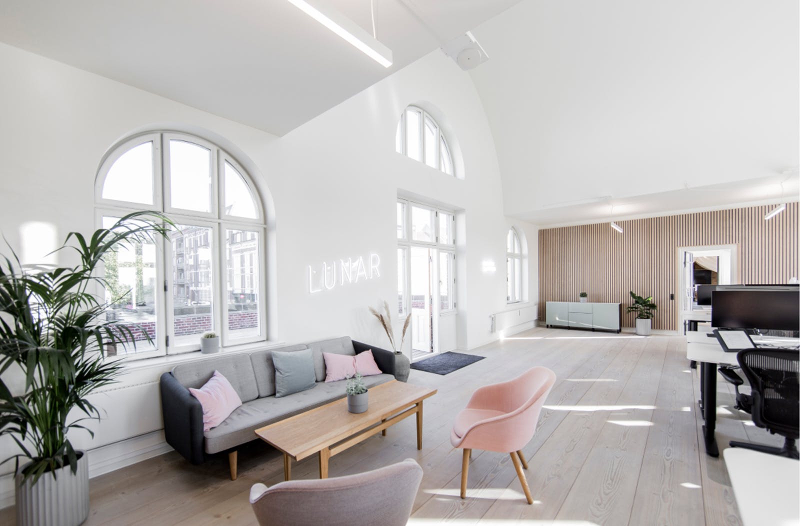 A bright and spacious office lounge area with high ceilings, arched windows, and a white neon "LUNAR" sign on the wall above a modern sofa.
