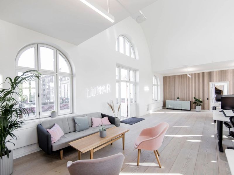 A bright and spacious office lounge area with high ceilings, arched windows, and a white neon "LUNAR" sign on the wall above a modern sofa.