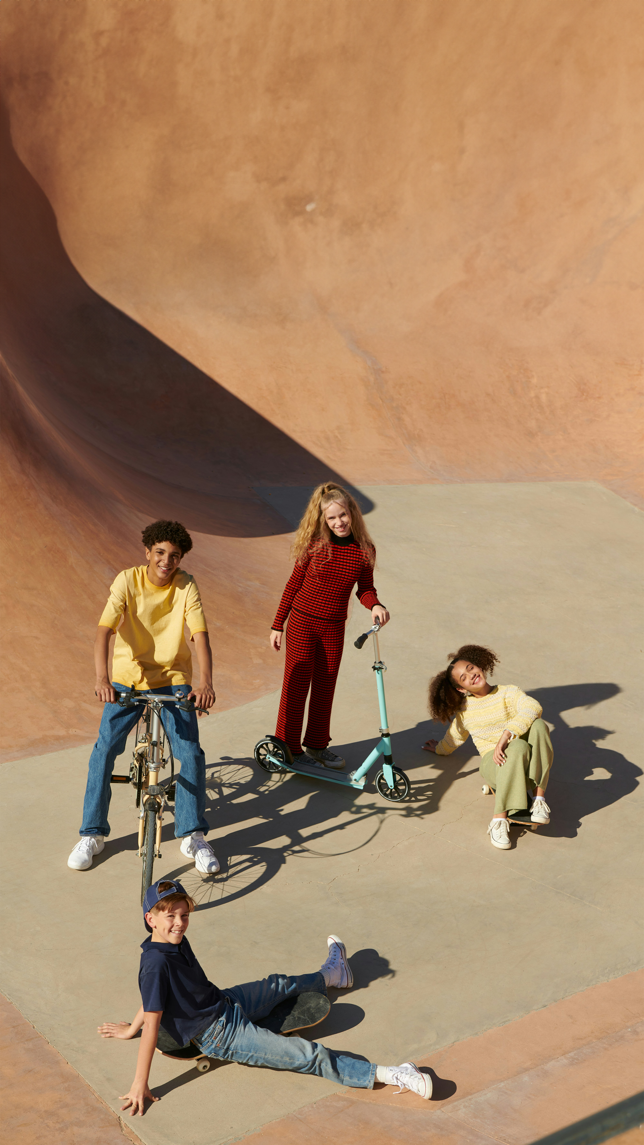 En mangfoldig gruppe på fire barn og tenåringer henger i en skatepark i betong med en sykkel, en sparkesykkel og et skateboard.