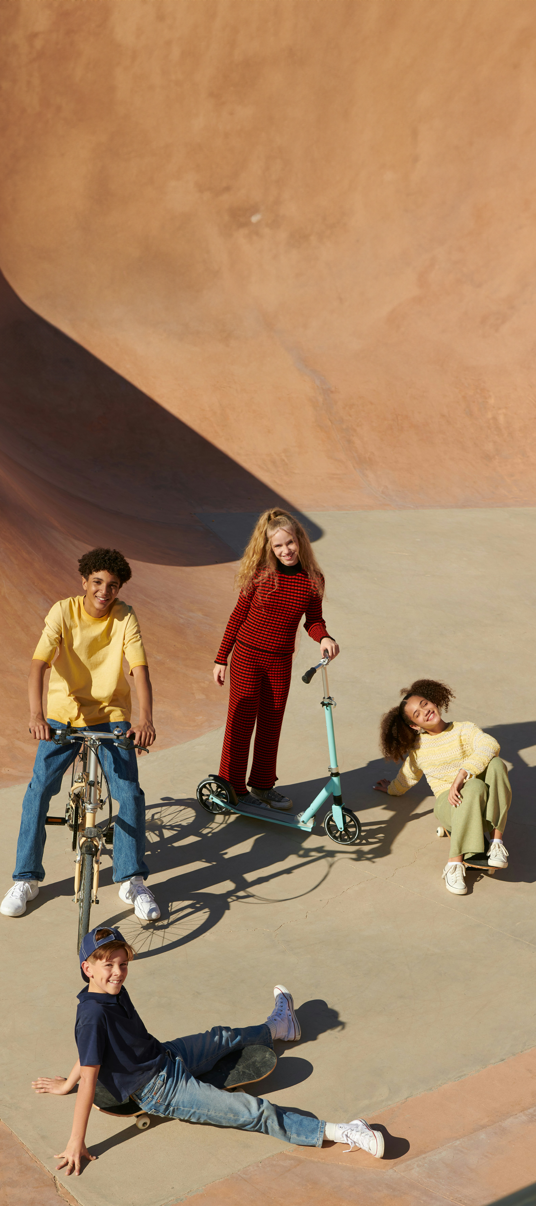 En mångfaldig grupp på fyra barn och tonåringar hänger i en skatepark i betong med en cykel, en sparkcykel och en skateboard.