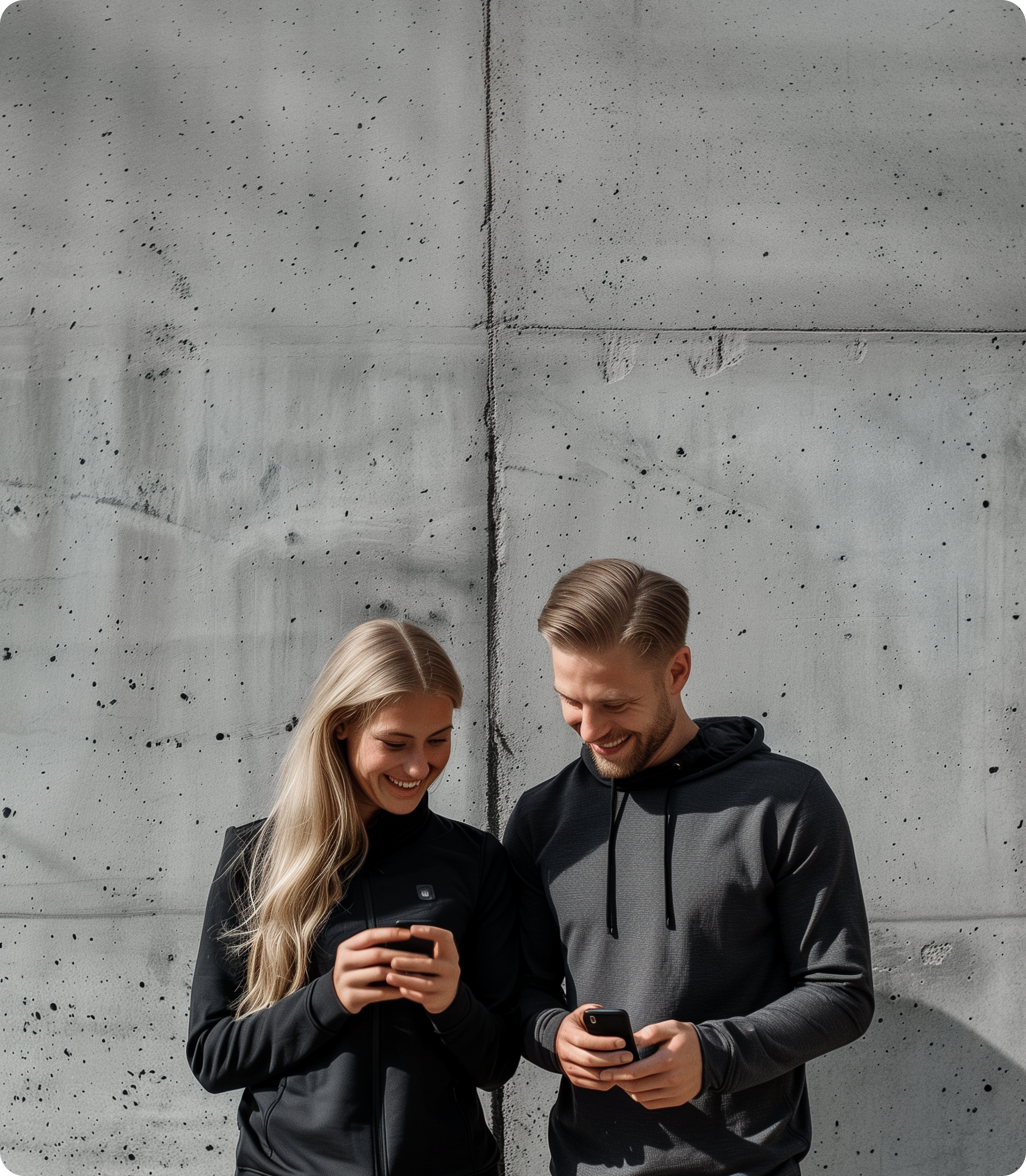 En mand og en kvinde i træningstøj står foran en betonvæg og smiler, mens de kigger ned på hver deres smartphone.