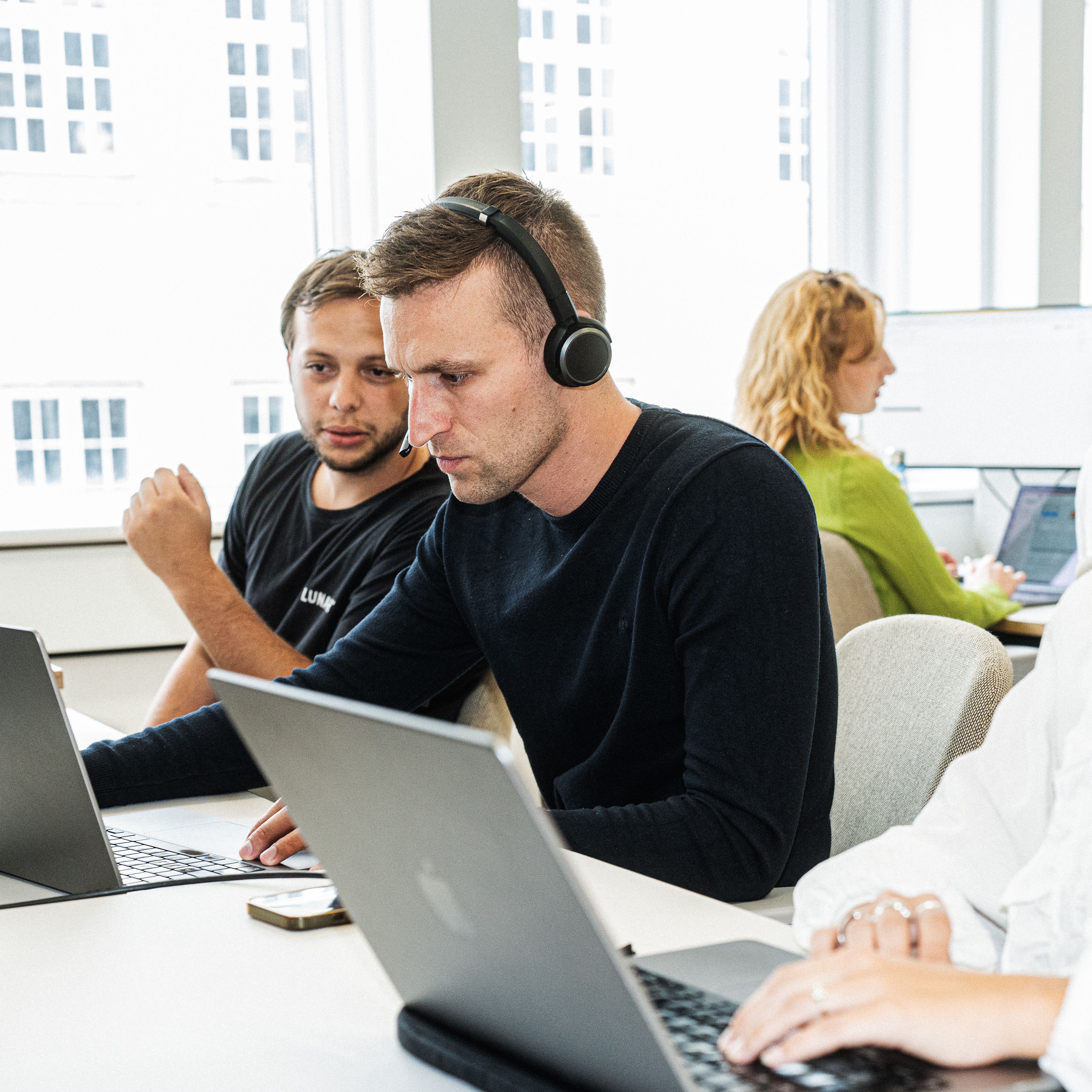 To mannlige kolleger samarbeider ved en pult i et moderne kontor. Mannen i forgrunnen har på seg et headset og fokuserer på laptopen sin, mens kollegaen hans ser på.