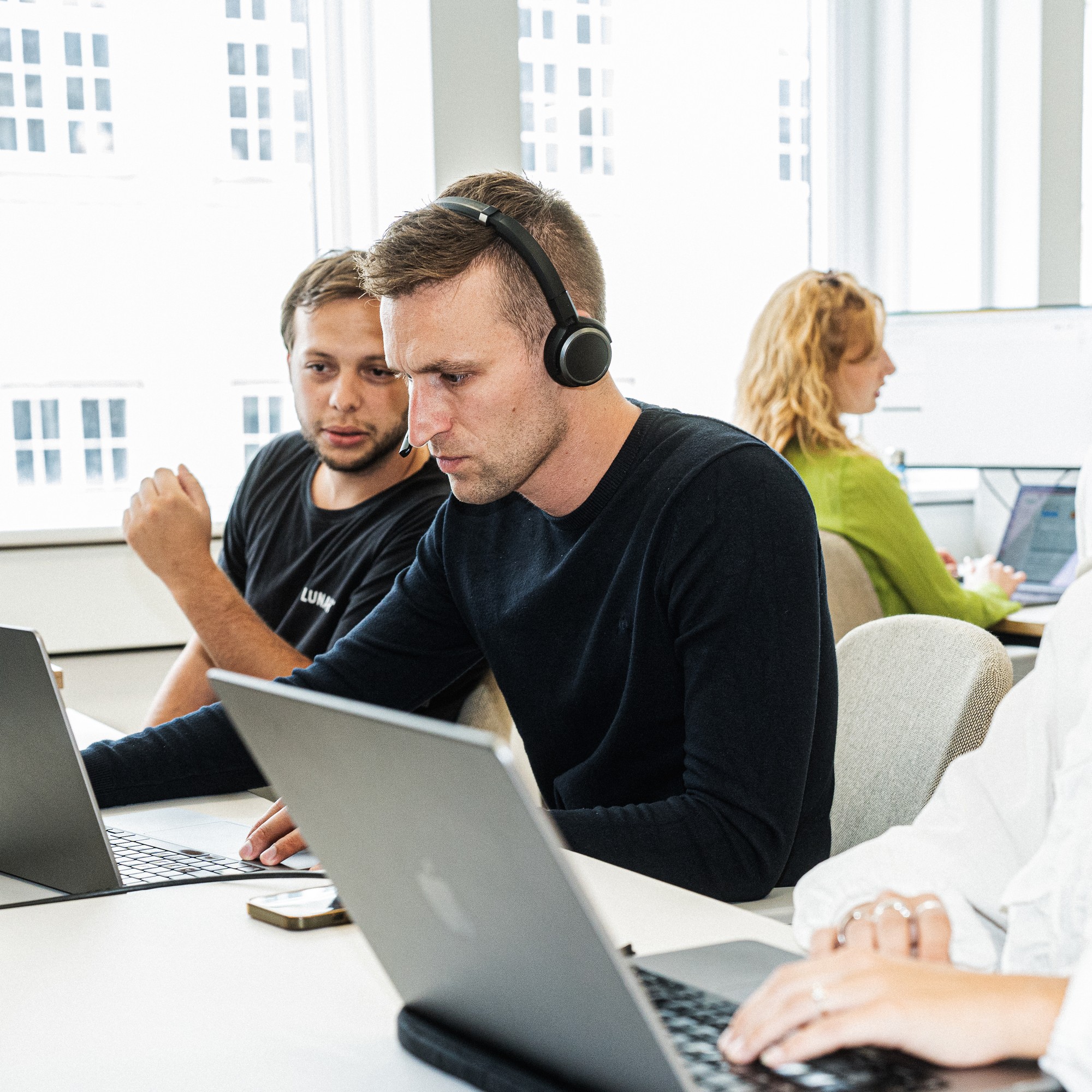 Två manliga kollegor samarbetar vid ett skrivbord på ett modernt kontor. Mannen i förgrunden bär ett headset och fokuserar på sin laptop, medan hans kollega ser på.