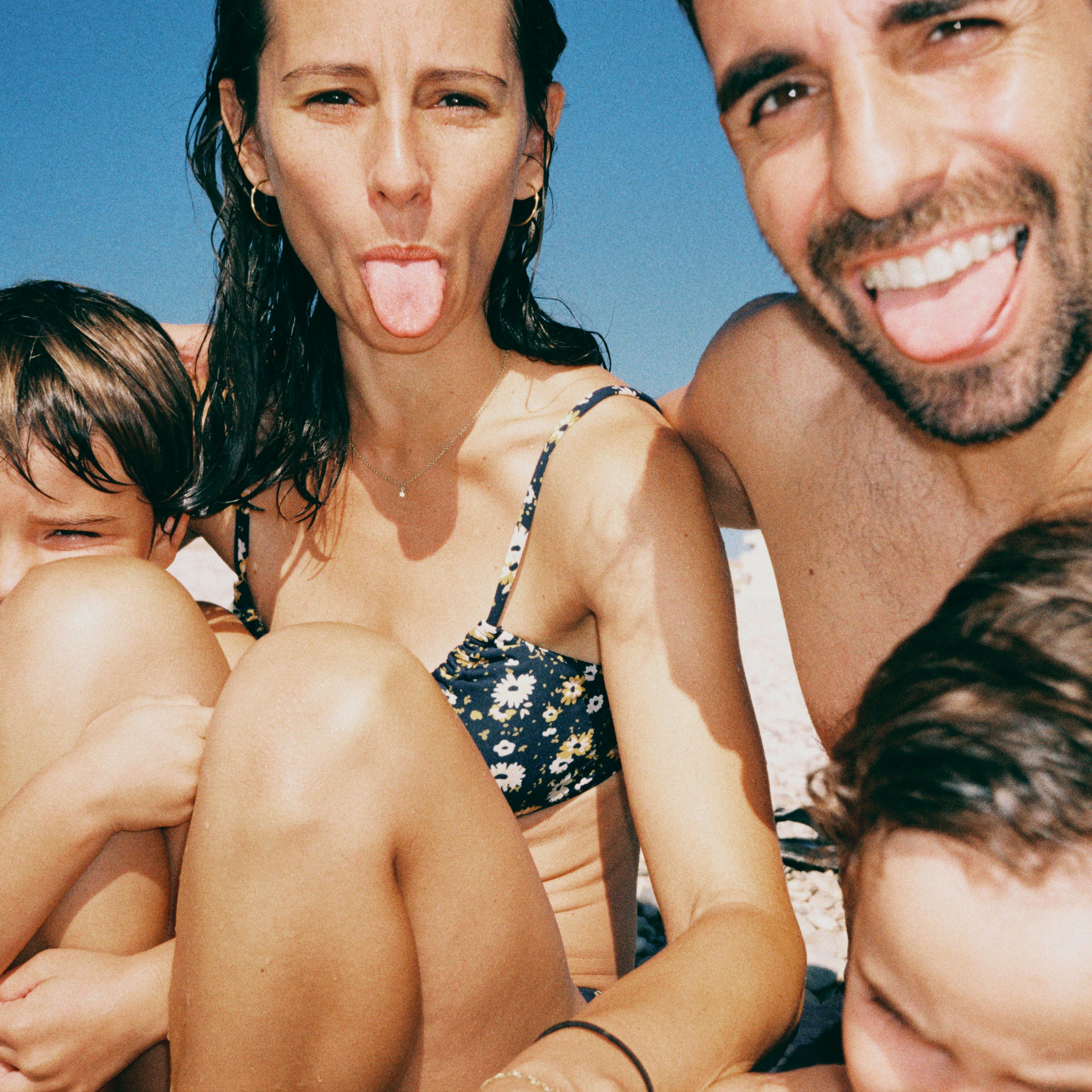  A close-up, playful family selfie at the beach. A man and woman smile and stick their tongues out at the camera, with their children partially in the shot.