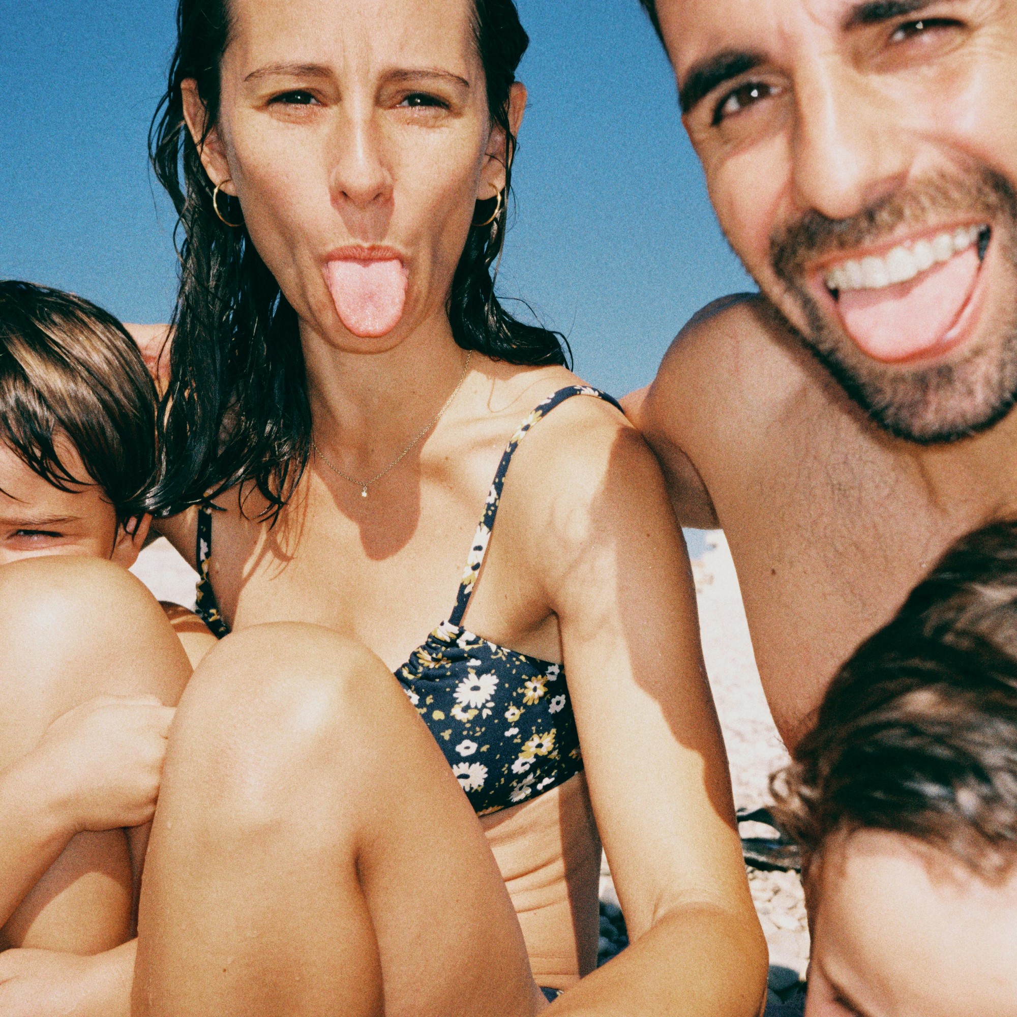 A close-up, playful family selfie at the beach. A man and woman smile and stick their tongues out at the camera, with their children partially in the shot.