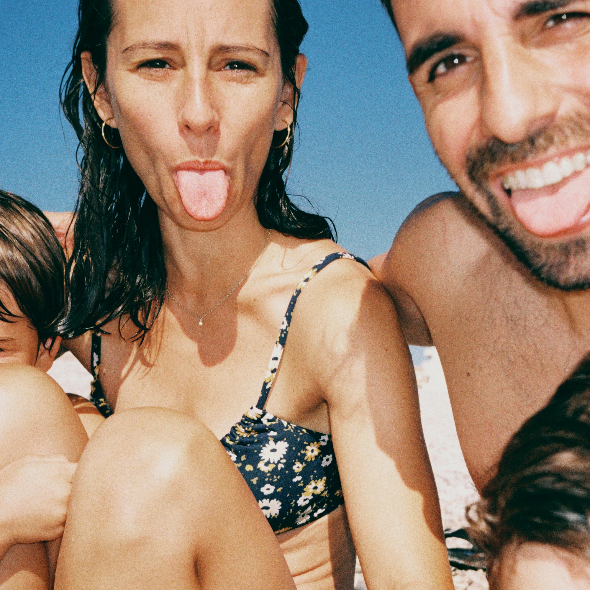 Et nærbilde av en familieselfie på stranden. En mann og en kvinne smiler og rekker tunge til kameraet, med barna sine delvis i bildet.