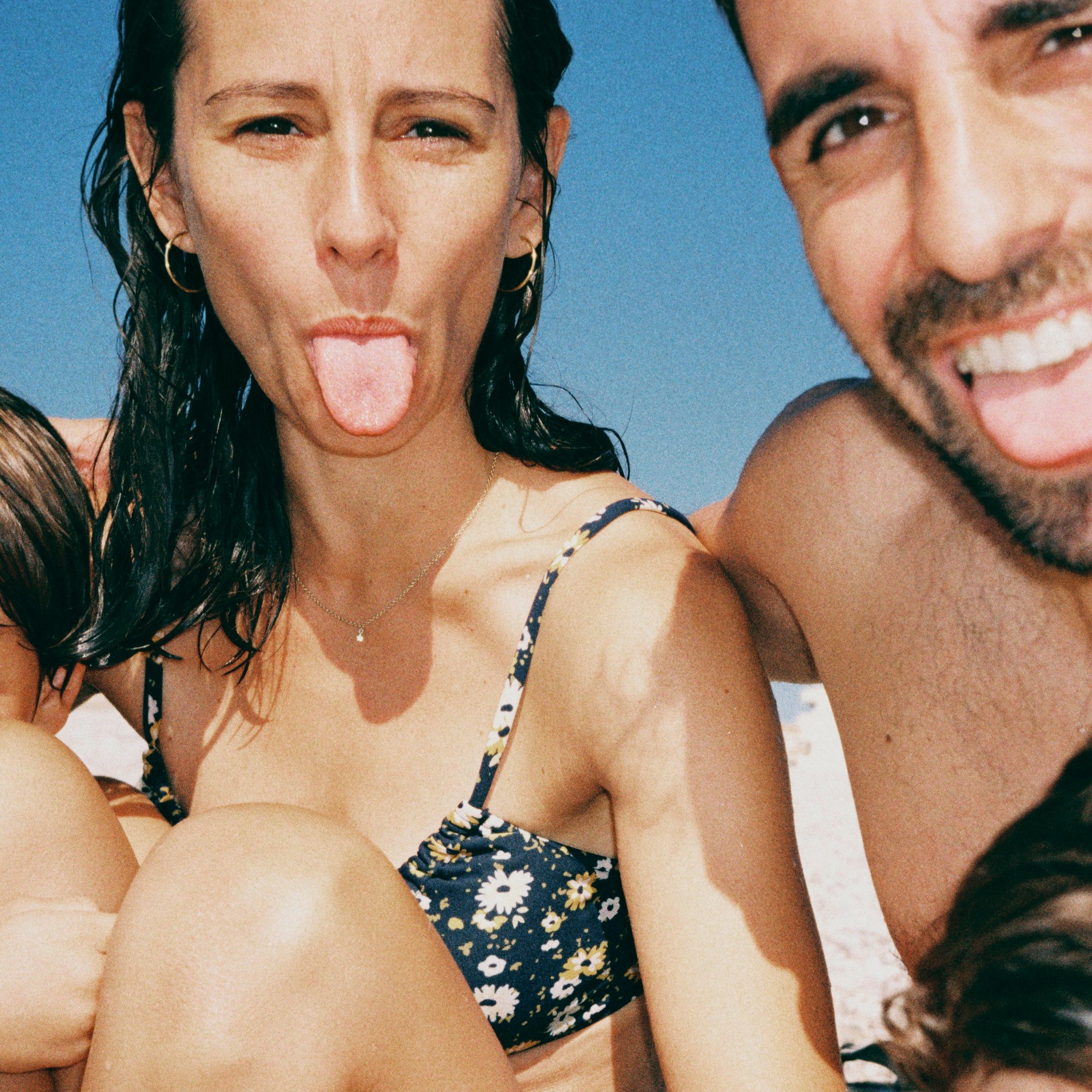 Et nærbillede af et familieselfie på stranden. En mand og en kvinde smiler og rækker tunge ad kameraet, mens deres børn er delvist med på billedet.